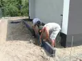 Dwóch mężczyzn pracuje nad przygotowaniem podłoża pod kostkę, układając obrzeża.