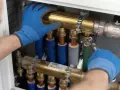 Ręce w niebieskich rękawiczkach dokręcają kluczem mosiężny element instalacji hydraulicznej w domu.
