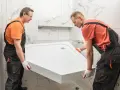 Dwóch fachowców montuje biały brodzik akrylowy bez nóżek w łazience z płytkami imitującymi marmur.