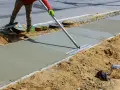 Robotnik wygładza świeży beton na chodniku. Prawdopodobnie używa mieszanki beton C20/25, której proporcje zapewniają trwałość.