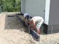 Dwóch mężczyzn układa suchy beton pod obrzeża wokół domu.