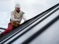 Dekarz w kasku i z pasem narzędziowym pracuje na dachu, przybijając młotkiem elementy pokrycia.
