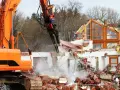 Pomarańczowa koparka niszczy budynek. Trwa wyburzenie, cena za m3 jest kluczowa przy takich pracach.