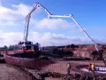 Ogromna pompa do betonu pracuje na budowie, dostarczając materiał do wykopu. Pracownicy w kamizelkach obserwują proces.