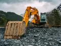 Żółta koparka na kamienistym zboczu, w tle las i ośnieżone góry. Zastanawiasz się, ile zarabia operator koparki w Norwegii?