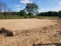 Przygotowanie terenu pod budowę: wyrównana piaszczysta działka z zaznaczonymi granicami, otoczona drzewami.