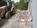 Żółta koparko-ładowarka pracuje przy wyburzeniu domu. Gruz i cegły leżą na ziemi. Koszt wyburzenia domu jest wysoki.