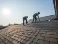 Dwóch dekarzy w kaskach i szelkach bezpieczeństwa pracuje na dachu. Jeden z nich używa wiertarki.