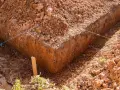 Głęboki wykop pod fundament, z widocznymi nićmi wyznaczającymi linie budowy. Widać błotniste dno i usypaną ziemię.