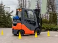Pomarańczowo-szary wózek widłowy Toyota z butlą gazową, otoczony żółtymi pachołkami. Zastanawiasz się, ile trwa kurs na wózek widłowy?