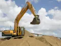 Żółta koparka Hyundai na placu budowy, z łyżką unoszącą się nad piaszczystym usypiskiem, tworzącym naturalny kąt stoku. W tle most i błękitne niebo z chmurami.