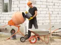 Budowlaniec wsypuje cement do betoniarki 100l. Obok stoi taczka i łopata.
