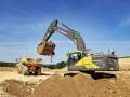 Koparka Volvo wysypuje ziemię do wywrotki. Trwa budowa, a pytanie "ile kosztuje metr wykopu pod kabel" jest kluczowe dla inwestycji.