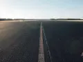 Asfaltowa droga z linią pośrodku, otoczona polami i lasem.