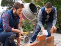 Dwóch budowlańców układa cegły. Jeden sprawdza poziom, drugi kładzie cegłę. W tle betoniarka, która pomoże ustalić, ile cementu na kubik betonu.