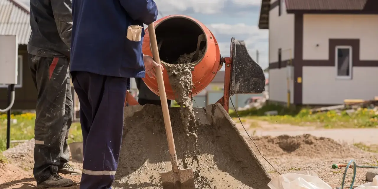 Dwóch budowlańców miesza beton w betoniarce. Widać, ile żwiru na worek cementu potrzeba do uzyskania odpowiedniej konsystencji.