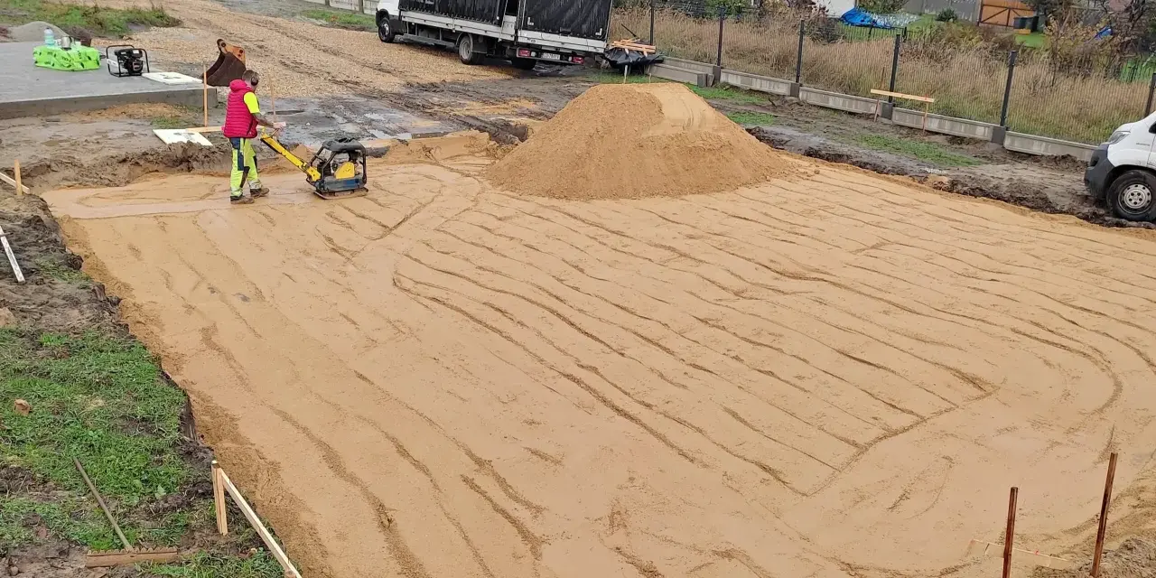 Pracownik zagęszcza piasek zagęszczarką. To kluczowy etap wymiany gruntu pod płytę fundamentową.