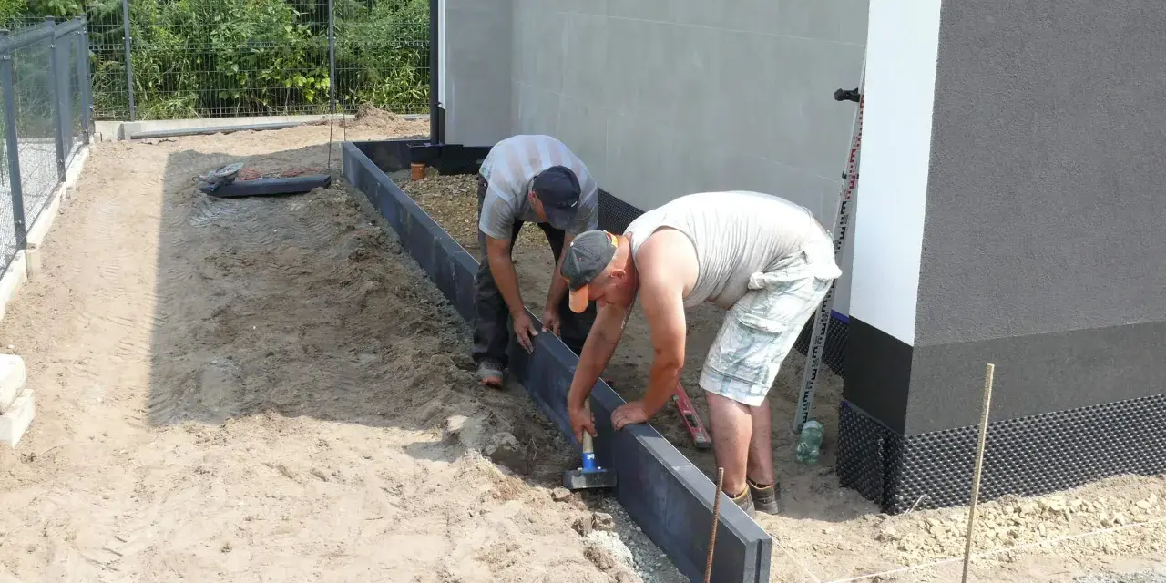 Dwóch mężczyzn układa suchy beton pod obrzeża wokół domu.