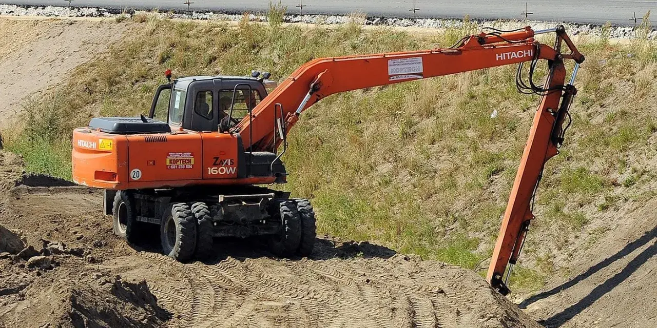 Pomarańczowa koparka Hitachi Zaxis 160W pracuje na budowie. Operator z uprawnieniami na minikoparkę wykonuje prace ziemne.