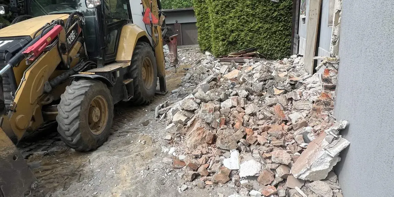 Koparka pracuje przy rozbiórce domu. Gruz i cegły leżą na ziemi, szacowany koszt rozbiórki domu jest wysoki.