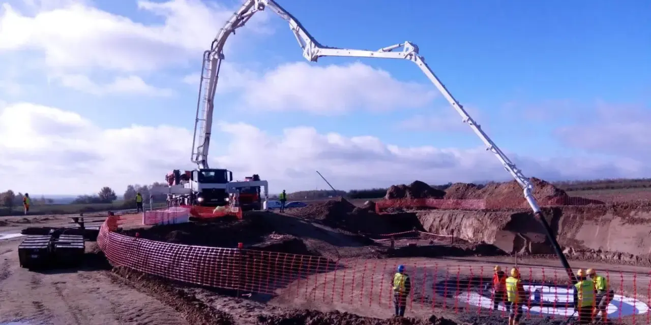 Ogromna pompa do betonu pracuje na budowie, dostarczając materiał do wykopu. Pracownicy w kamizelkach obserwują proces.