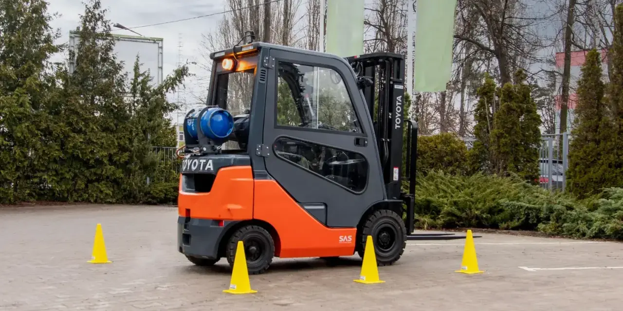 Pomarańczowo-szary wózek widłowy Toyota z butlą gazową, otoczony żółtymi pachołkami. Zastanawiasz się, ile trwa kurs na wózek widłowy?