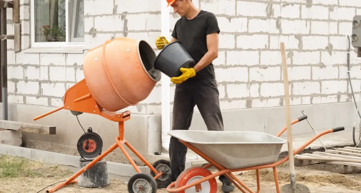 Budowlaniec wsypuje cement do betoniarki 100l. Obok stoi taczka i łopata.