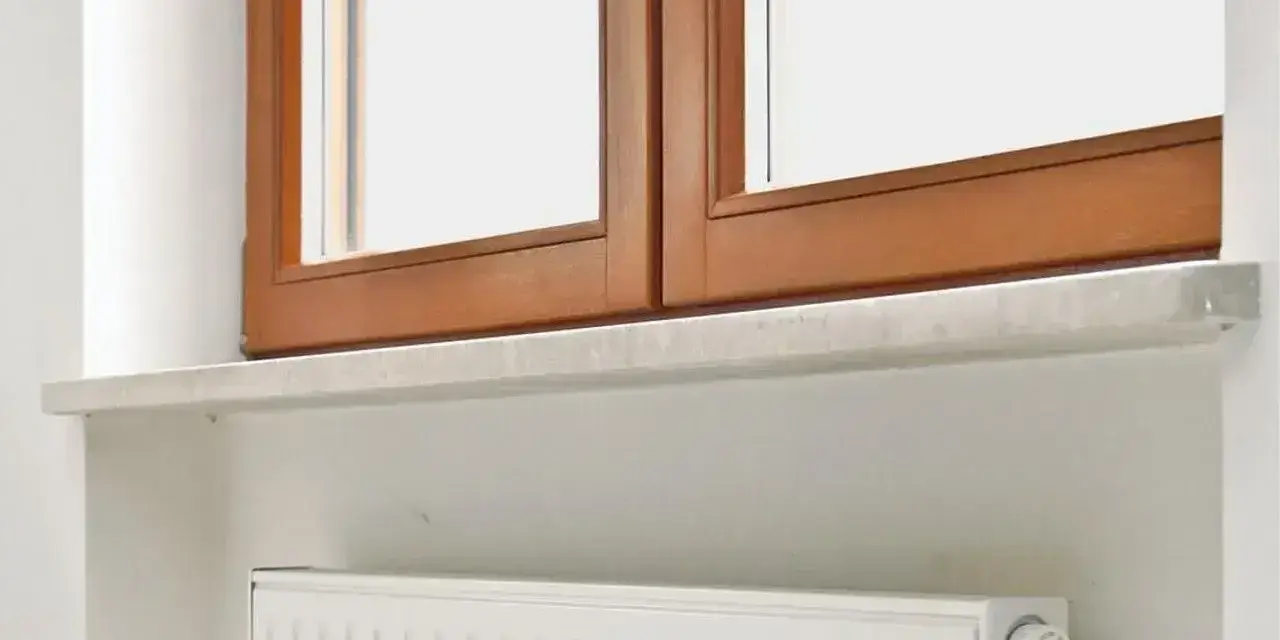 Elegancki montaż parapetów wewnętrznych cena. Biały grzejnik pod drewnianym oknem.