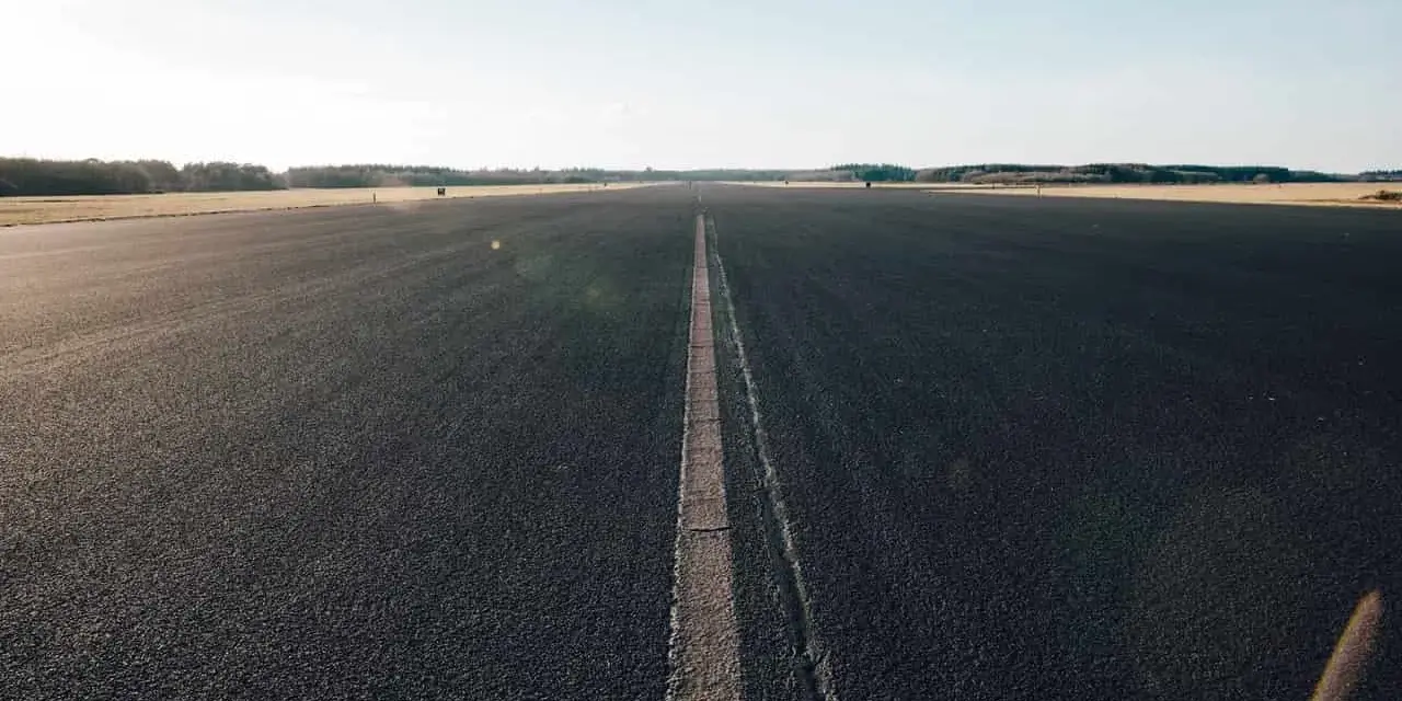 Asfaltowa droga z linią pośrodku, otoczona polami i lasem.