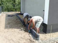 Dwóch mężczyzn pracuje nad przygotowaniem podłoża pod kostkę, układając obrzeża.