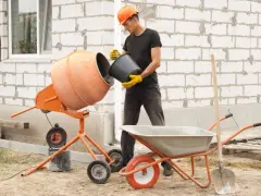 Budowlaniec wsypuje cement do betoniarki 100l. Obok stoi taczka i łopata.