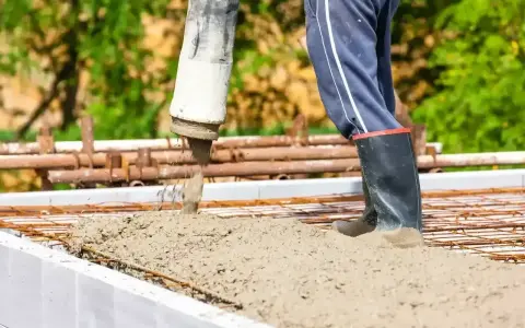 Beton wylewany do szalunków fundamentowych. Pracownik w gumowcach nadzoruje proces.