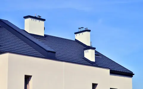Nowoczesny dach bezokapowy z ciemnogranatową dachówką i białymi kominami, pod błękitnym niebem.