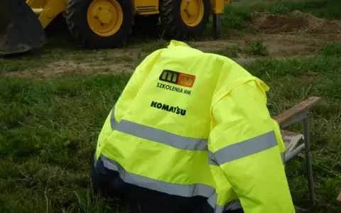 Żółta kamizelka z logo Komatsu i napisem "Szkolenia MM" leży na krześle. To przygotowanie do egzaminu na ładowarkę, gdzie pytania i odpowiedzi są kluczowe.