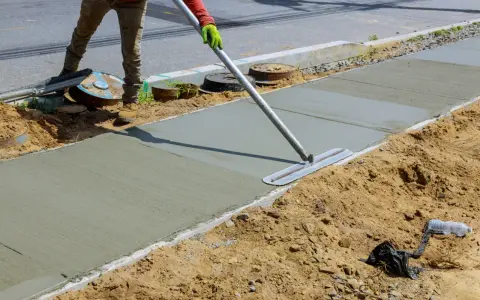 Robotnik wygładza świeży beton na chodniku. Prawdopodobnie używa mieszanki beton C20/25, której proporcje zapewniają trwałość.