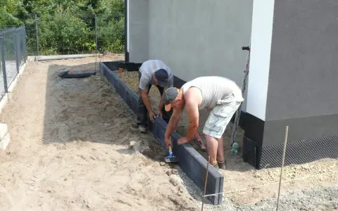 Dwóch mężczyzn układa suchy beton pod obrzeża wokół domu.