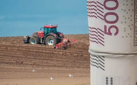 Czerwony traktor orze pole, a na pierwszym planie zwinięte banknoty 500 zł. Zastanawiasz się, ile kosztuje hektar ziemi budowlanej?