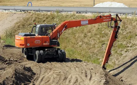 Pomarańczowa koparka Hitachi Zaxis 160W pracuje na budowie. Operator z uprawnieniami na minikoparkę wykonuje prace ziemne.