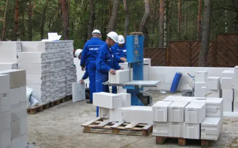 Budowa trwa! Pracownicy w niebieskich kombinezonach i białych kaskach układają bloczki ytong. Na paletach widać, że ytong 12 to spora ilość materiału budowlanego.