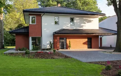 Nowoczesny dom z ceglanymi akcentami i eleganckim, czarnym dachem czterospadowym, otoczony zielenią.