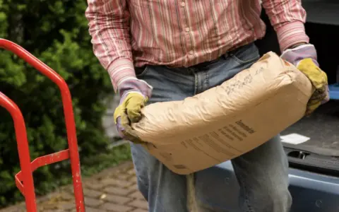 Mężczyzna w rękawicach niesie worek cementu, zastanawiając się, ile cementu do piasku potrzeba do zaprawy.