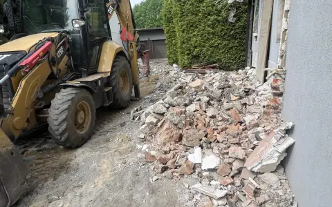 Koparka pracuje przy rozbiórce domu. Gruz i cegły leżą na ziemi, szacowany koszt rozbiórki domu jest wysoki.