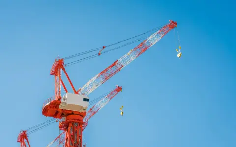 Dwa czerwono-białe żurawie budowlane na tle błękitnego nieba. Widać drabinki i platformy, które pokazują, jak się wchodzi na żurawia.