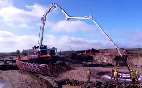 Ogromna pompa do betonu pracuje na budowie, dostarczając materiał do wykopu. Pracownicy w kamizelkach obserwują proces.