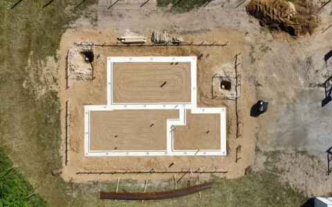 Widok z lotu ptaka na plac budowy. Widać fundamenty domu, piasek, drewno i wykopy pod studnie fundamentowe.
