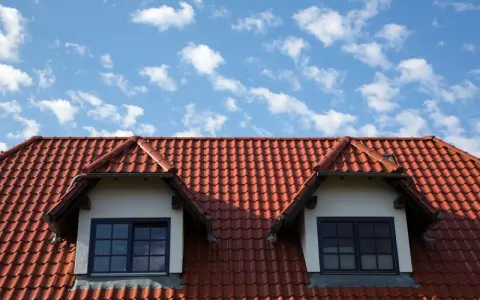 Czerwony dach z dachówki ceramicznej, z dwoma lukarnami i niebieskim niebem w chmurach.