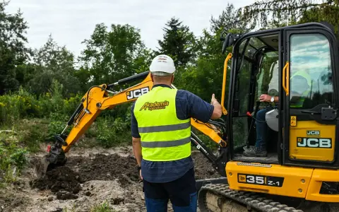 Instruktor nadzoruje kurs koparki klasa 3. Operator w kabinie, drugi pracownik w kamizelce odblaskowej.