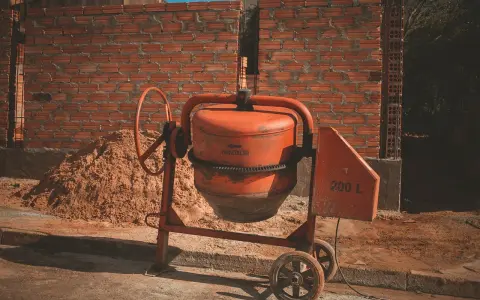 Pomarańczowa betoniarka na budowie, obok sterty piasku i muru z cegieł.