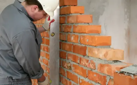 Murarz w kasku sprawdza pion muru, układając cegły. Widać precyzję w dopasowaniu wymiarów cegły do budowanej ściany.