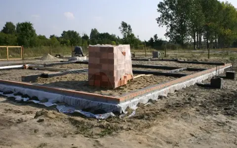 Budowa domu w toku. Widać zarys fundamentów, zbrojenie i ułożoną cegłę. Na środku placu budowy stoi paleta cegieł.